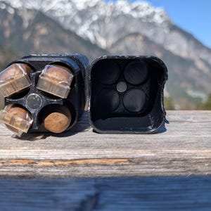May include: Black, open container with four compartments, each holding a small, round object. The container is next to a matching, empty container. The background features a wooden surface and a mountain range.