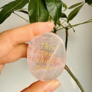 Puede incluir: Una piedra de cuarzo rosa con el texto dorado "This too shall pass". La piedra ovalada se sostiene frente a una planta verde. La piedra tiene un tono rosa translúcido.