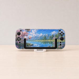May include: A handheld gaming console featuring a decorative skin with a landscape illustration of a blue lake, green pine trees, snow-capped mountains, and pink cherry blossom branches under a bright blue sky with white clouds.