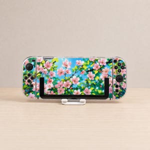 May include: A handheld gaming console featuring a decorative skin with a vibrant pattern of pink cherry blossoms against a bright blue sky. The device includes black control buttons, joysticks, and a directional pad, all mounted on a white stand.