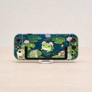 May include: A handheld gaming console featuring a clear protective case with a printed design of green frogs, pink lotus flowers, lily pads, and a boba tea drink on a dark blue water background, held in a black and white plastic display stand.