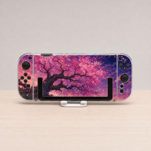 May include: A handheld gaming console featuring a vibrant skin with a dark tree silhouette against a bright pink and purple cherry blossom sky. The device has black buttons, joysticks, and a directional pad, and it is mounted on a white stand.