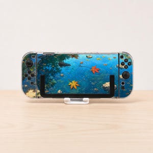 May include: A handheld gaming console featuring a clear protective case and a decorative skin displaying a vibrant blue pond scene with floating orange and yellow maple leaves, resting on a white stand against a plain light background.