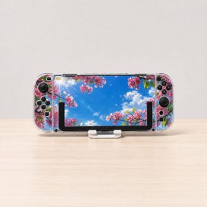 May include: A handheld gaming console featuring a clear protective case with a printed design of pink cherry blossom branches against a bright blue sky with white clouds and sun rays, held in a white plastic stand on a light wooden surface.