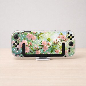 May include: A handheld gaming console featuring a clear protective case with a printed design of pink cherry blossoms on branches against a soft green and white bokeh background. The device includes black joysticks, buttons, and a black stand.