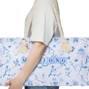 Puede incluir: Un bolso tote rectangular con base blanca, estampado floral azul, p&aacute;jaros y una ilustraci&oacute;n de una pagoda. La palabra MAHJONG est&aacute; impresa en letras azules en el centro. El bolso tiene asas de cuerda gruesa de color crema con nudos.