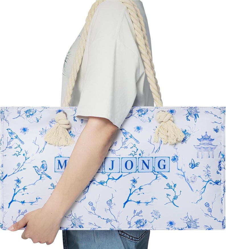 Puede incluir: Un bolso tote rectangular con base blanca, estampado floral azul, p&aacute;jaros y una ilustraci&oacute;n de una pagoda. La palabra MAHJONG est&aacute; impresa en letras azules en el centro. El bolso tiene asas de cuerda gruesa de color crema con nudos.