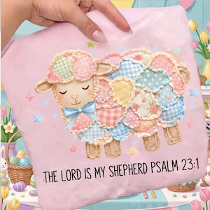 May include: A light pink t-shirt featuring a colorful patchwork sheep design with a blue bow. The text "THE LORD IS MY SHEPHERD PSALM 23:1" is printed in black. The sheep design includes various pastel colors and star accents.