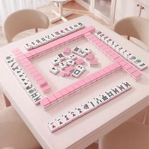 Może przedstawiać: Zestaw do gry w Mahjong składający się z białych płytek z czarnymi i czerwonymi znakami oraz r&oacute;żowymi rewersami. Płytki są ułożone w kwadratowy obw&oacute;d na stole, a w środku pola gry znajdują się dodatkowe płytki oraz dwie małe r&oacute;żowe kostki.
