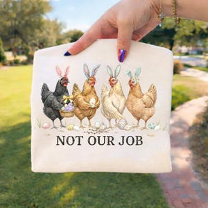 May include: A beige tote bag featuring a whimsical illustration of four chickens wearing bunny ears and holding Easter eggs. The text "NOT OUR JOB" is printed below the chickens. The bag is held by a person.
