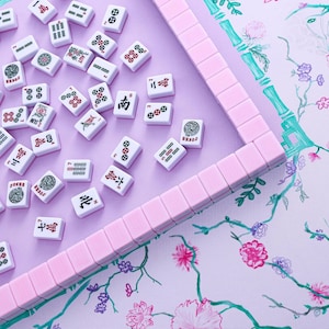 Puede incluir: Un juego de mahjong con fichas blancas que presentan s&iacute;mbolos, n&uacute;meros y caracteres en rojo, verde y negro, esparcidas sobre una superficie morada clara. Una fila de fichas rosa claro forma un marco parcial sobre un fondo con motivos florales y de bamb&uacute;.