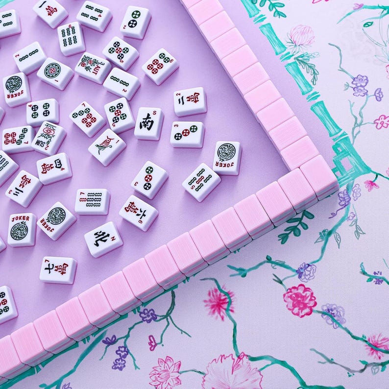 Puede incluir: Un juego de mahjong con fichas blancas que presentan s&iacute;mbolos, n&uacute;meros y caracteres en rojo, verde y negro, esparcidas sobre una superficie morada clara. Una fila de fichas rosa claro forma un marco parcial sobre un fondo con motivos florales y de bamb&uacute;.