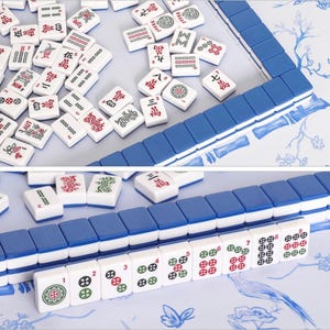 Puede incluir: Un conjunto de fichas de mahjong blancas con el reverso azul, que presentan varios s&iacute;mbolos, caracteres y n&uacute;meros grabados en rojo, verde y negro. Algunas fichas est&aacute;n dispuestas en filas ordenadas, mientras que otras est&aacute;n esparcidas sobre una superficie con motivos florales azules.