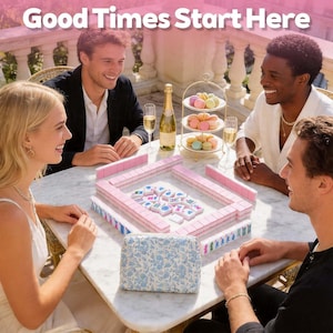 May include: A mahjong set with pink tiles arranged on a marble table, featuring a blue and white floral patterned storage bag, a three-tiered dessert stand with colorful macarons, and a bottle of champagne with two glasses, text reads Good Times Start Here.