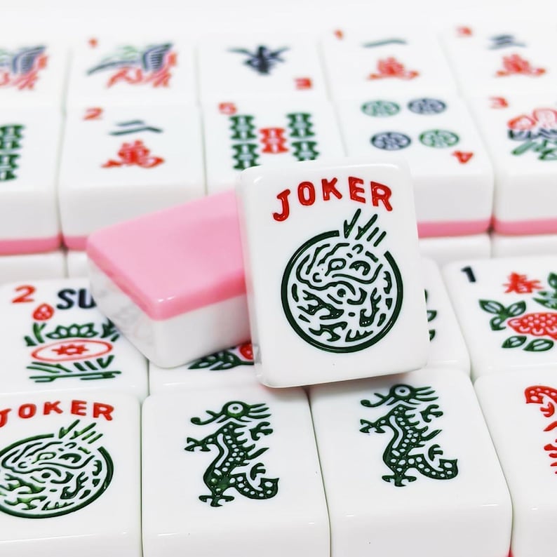 Może przedstawiać: Kolekcja białych płytek do gry w Mahjong z r&oacute;żnymi wygrawerowanymi zielonymi i czerwonymi symbolami, w tym motywami smok&oacute;w oraz napisem JOKER w kolorze czerwonym. Jedna płytka stoi pionowo, ukazując okrągły wz&oacute;r oraz czerwony napis JOKER.