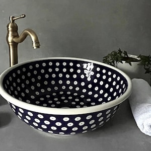 Puede incluir: Un lavabo azul marino con lunares blancos, un grifo de latón, un vaso, un jabonera y un dispensador de jabón a juego. También hay una toalla blanca enrollada y una pequeña planta.