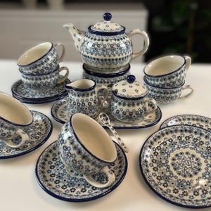 Op de afbeelding: Een complete theeset met een bloemenpatroon in blauw- en wittinten. De set bevat een theepot, kopjes, schotels, een melkkan, een suikerpot en lepels. De theeset is gerangschikt op een wit oppervlak.