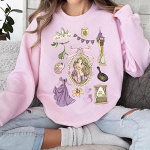 May include: Pink sweatshirt featuring a collection of illustrations from a fairytale. The design includes a portrait of a princess, a tower, a frying pan, a dress, and other related elements. The sweatshirt is a light pink color.