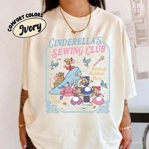 May include: Ivory-colored t-shirt featuring a graphic of Cinderella's Sewing Club. The design includes Cinderella's glass slipper, mice, and the text "Bibbidi Bobbidi Boo." The shirt also has the text "Est. 1950" and "Comfort Colors" on the top left.