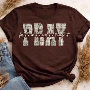 May include: A dark brown t-shirt with the words "PRAY" and "for it. on it. over it. through it" printed in a distressed, newspaper-like font. The shirt is paired with blue jeans and dried ornamental grass.
