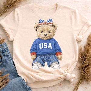 Peut inclure: T-shirt crème avec un ours en peluche portant un pull bleu avec "USA" imprimé, un jean bleu et des baskets blanches. L'ours a un nœud avec un motif étoiles et rayures. Le t-shirt est noué en bas.