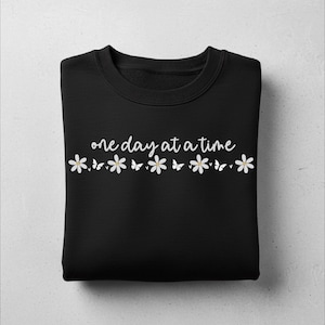 Peut inclure: Sweat-shirt noir avec l'inscription "one day at a time" en blanc. Une rangée de marguerites blanches et de papillons orne le bas du texte. Le vêtement est plié sur une surface gris clair.