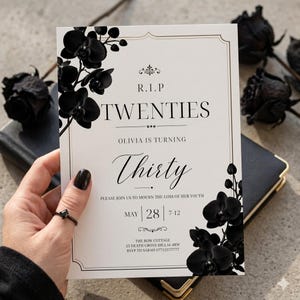 May include: A white invitation card with black text and floral accents. The card reads "R.I.P Twenties, Olivia is turning Thirty." Black orchid flowers frame the text. The date and time of the event are also included.