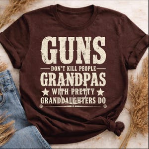 May include: A brown t-shirt with the text "GUNS DON'T KILL PEOPLE GRANDPAS WITH PRETTY GRANDDAUGHTERS DO" in distressed white lettering. The shirt is folded with a pair of blue jeans and dried grass.