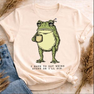 Peut inclure: T-shirt crème avec une illustration d'une grenouille verte tenant une tasse. La grenouille a une expression fatiguée et est soulignée en noir. Le texte "I HAVE TO SAY WEIRD STUFF OR I'LL DIE" est imprimé en noir.