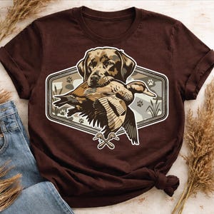 Op de afbeelding: Bruin t-shirt met een afbeelding van een Labrador Retriever die een eend draagt. De afbeelding staat in een zeshoekige lijst met een camouflage achtergrond en pootafdrukken.