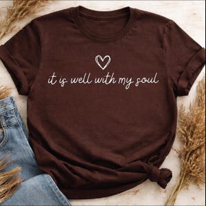 以下が含まれることがあります： 袖をまくった濃い茶色のTシャツ。白い筆記体で「it is well with my soul」というフレーズが書かれており、その上に小さなハートが描かれています。Tシャツは青いジーンズと組み合わせて表示されています。