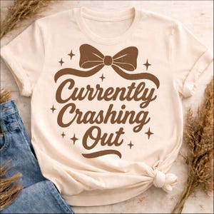 May include: Cream-colored t-shirt with a brown bow graphic and the text "Currently Crashing Out." The design includes star accents. The shirt is knotted at the bottom and paired with blue jeans.