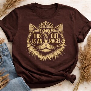 Peut inclure: T-shirt marron avec un motif de chat portant des lunettes et une couronne. Le texte sur le t-shirt se lit "THIS IS AN OUTRAGE!" dans une police circulaire. Le motif du chat est de couleur or.