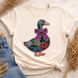 Peut inclure: T-shirt crème avec un motif de canard patchwork coloré. Le canard a une tête bleue et verte, un nœud papillon violet et un corps fait de divers tissus à motifs. Le canard est bordé de petites perles.