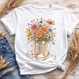 Peut inclure: T-shirt blanc avec un motif floral coloré. Un bouquet de fleurs éclatantes sort d'une paire de baskets montantes beiges. Les fleurs comprennent des fleurs orange, roses, bleues et jaunes, avec un papillon. Les baskets ont des détails rouges.