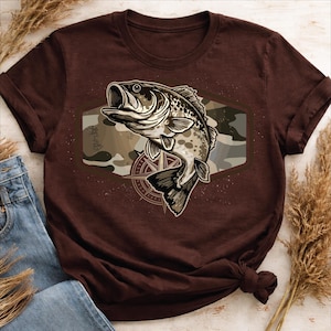 Pode incluir: Uma camiseta marrom com uma ilustração de um baixo saltando. O peixe está sobre um fundo de camuflagem dentro de uma forma hexagonal, com um desenho de rosa dos ventos abaixo. A camiseta está dobrada, com um par de jeans azuis visíveis.