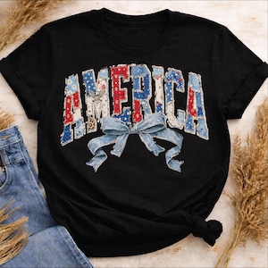 Peut inclure: T-shirt noir avec le mot "AMERICA" dans un motif patchwork rouge, blanc et bleu. Un nœud bleu clair est centré en dessous. Le t-shirt est associé à un jean bleu.