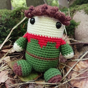 May include: A handmade crochet doll with a whimsical design. The doll features a cream-colored head with black button eyes and brown hair. It wears a green body with red accents and brown feet, sitting among fallen leaves.