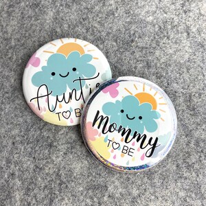 May include: Two round buttons with a colorful cloud and sun design. One button says "Auntie to Be" and the other says "Mommy to Be".