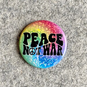 May include: A rainbow glitter button with the text "Peace Not War" in black, featuring a peace sign.