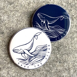 May include: Two round, blue and white enamel pins with a whale illustration and the text "Save the Ocean".