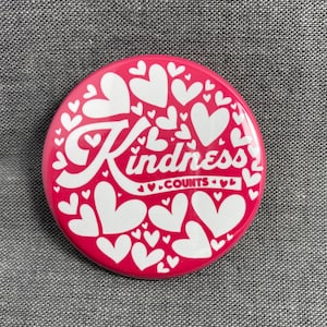 May include: A pink button with white hearts and the text "Kindness Counts" in a white stylized font.