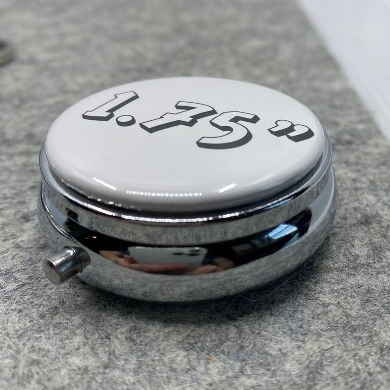 K&ouml;nnte beinhalten: Eine kleine, runde, silberne Pillendose mit einem wei&szlig;en Deckel. Der Deckel tr&auml;gt die Nummer "1.75" in Schwarz.