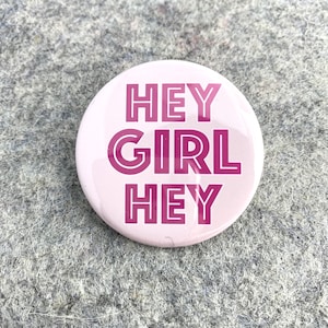 May include: A round, light pink button with the words "HEY GIRL HEY" in a darker pink, outlined font. The button is centered and set against a gray, textured background.