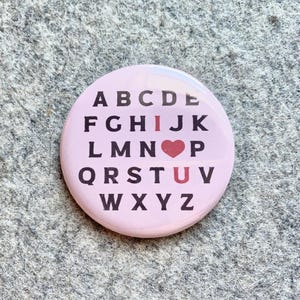 May include: A pink button with the alphabet printed in black letters. The letter "I" is replaced with a red heart and the letter "U" is highlighted in red. The text reads "I love you".