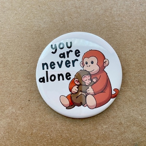 May include: A white button with the text "you are never alone" in black. The button features a cartoon illustration of a large orange monkey hugging a smaller brown monkey.