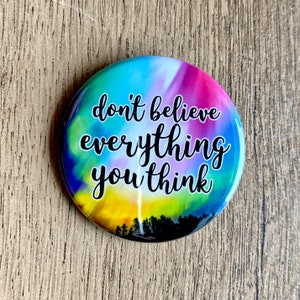 May include: A round button with a colorful tie-dye background and the text "don't believe everything you think" in black.