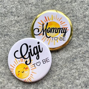 May include: Two round buttons with a sun design and text. The first button says "Mommy to be" and the second button says "Gigi to be".