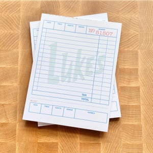 May include: Stack of white restaurant guest check pads with blue grid lines and text. The word "Lukes" is printed in light blue. The top check has the number "51507" printed in red.