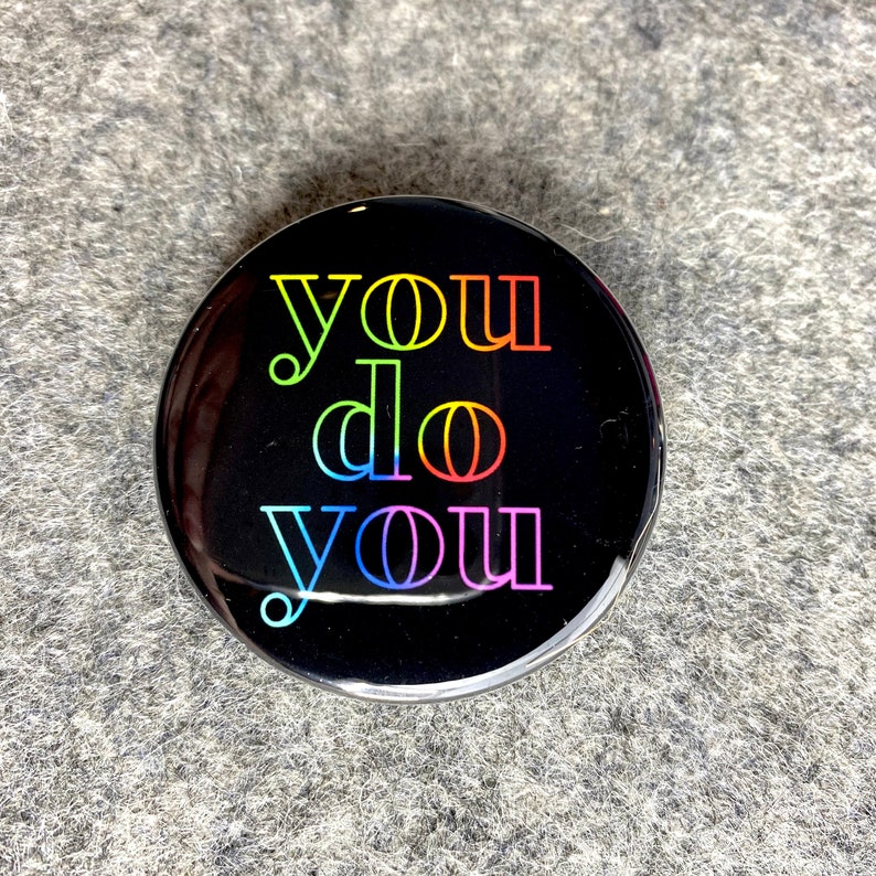K&ouml;nnte beinhalten: Ein schwarzer Knopf mit dem Text "you do you" in Regenbogenfarben.